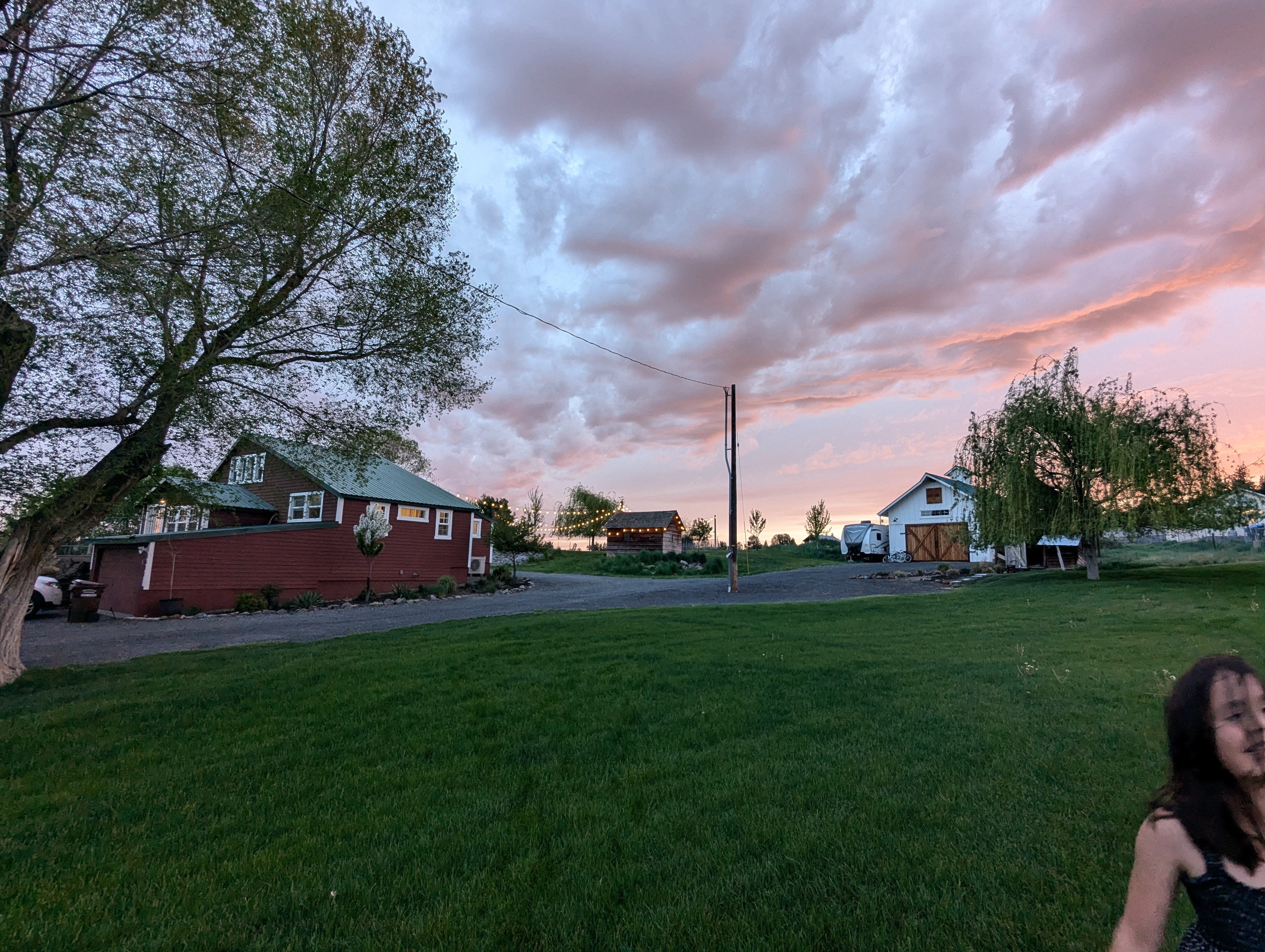 A picturesque rural scene featuring a red house with green accents, a white barn, and lush green grass under a colorful sunset sky with clouds.