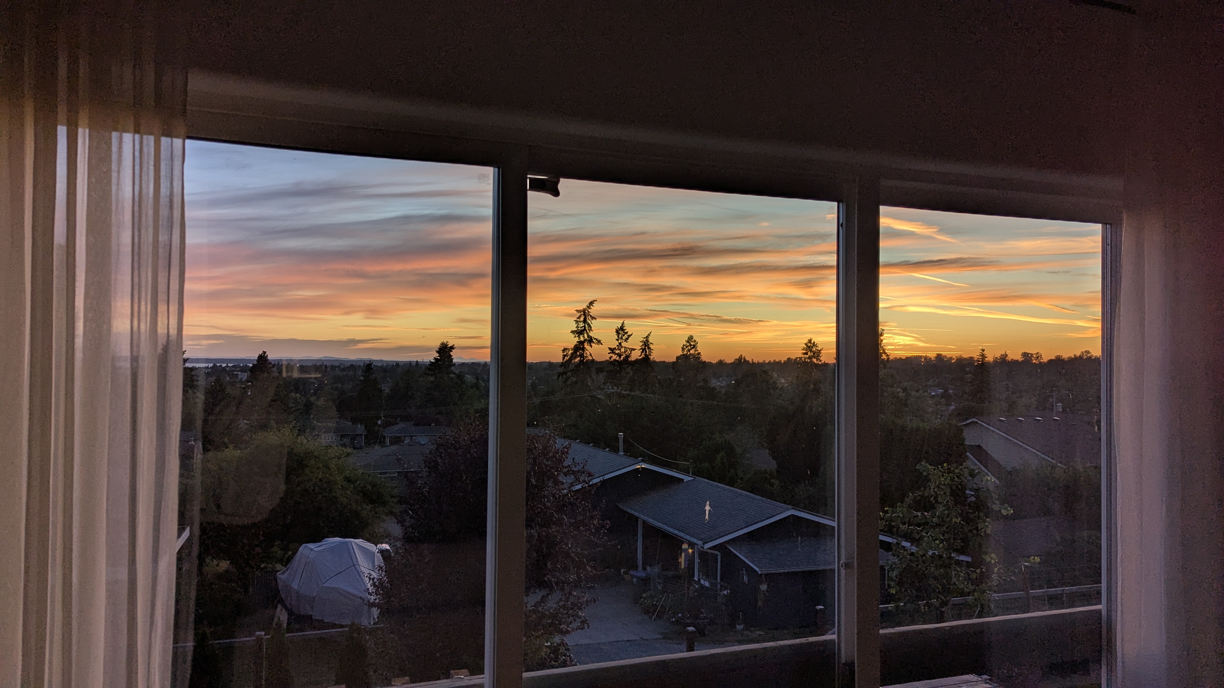 A beautiful sunset viewed through large windows, with vibrant orange and purple hues in the sky, silhouetted trees, and houses visible in the foreground.