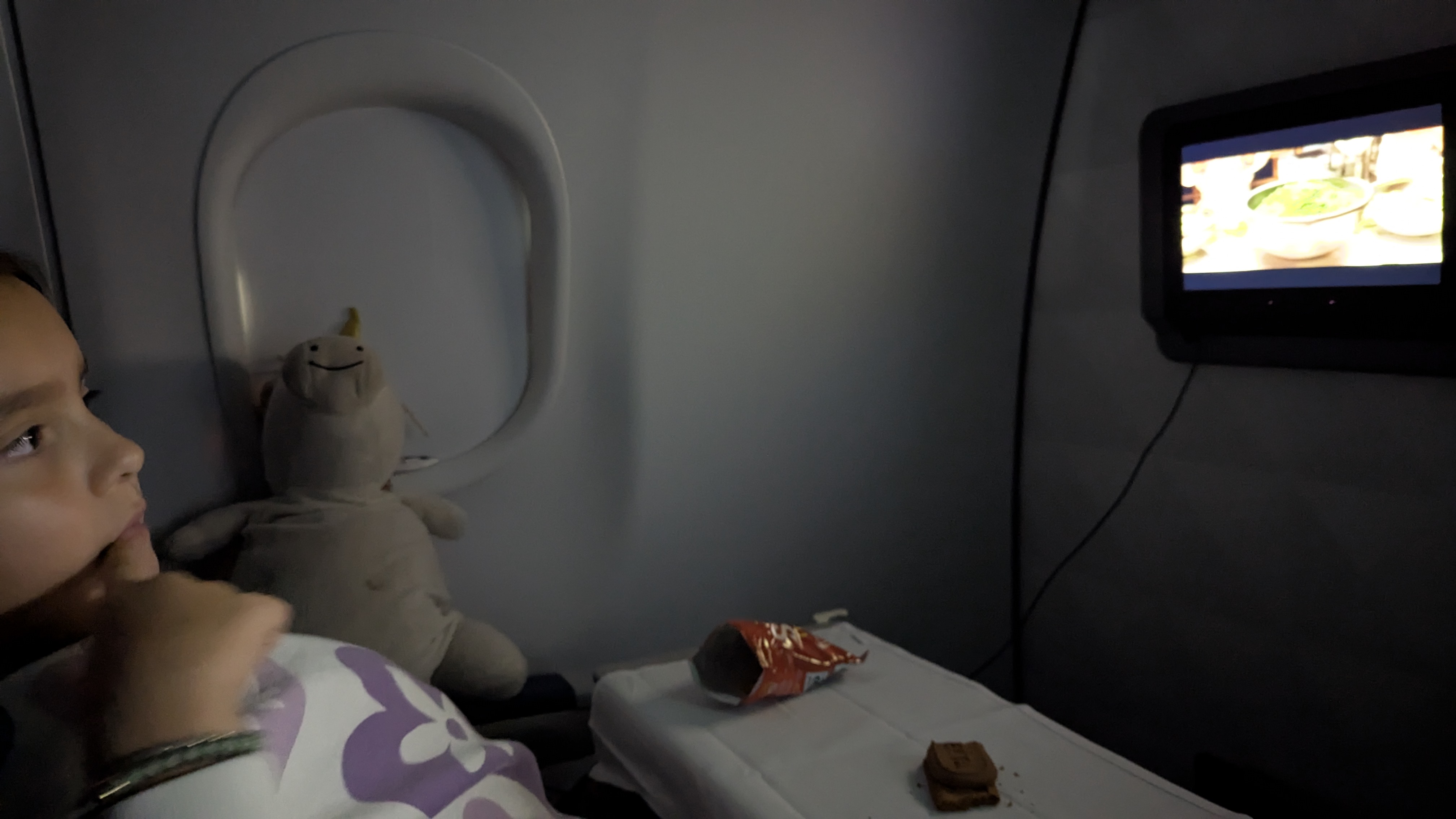 A child watching a screen in an airplane, with a stuffed toy beside them, snacks on the table, and a window in the background.