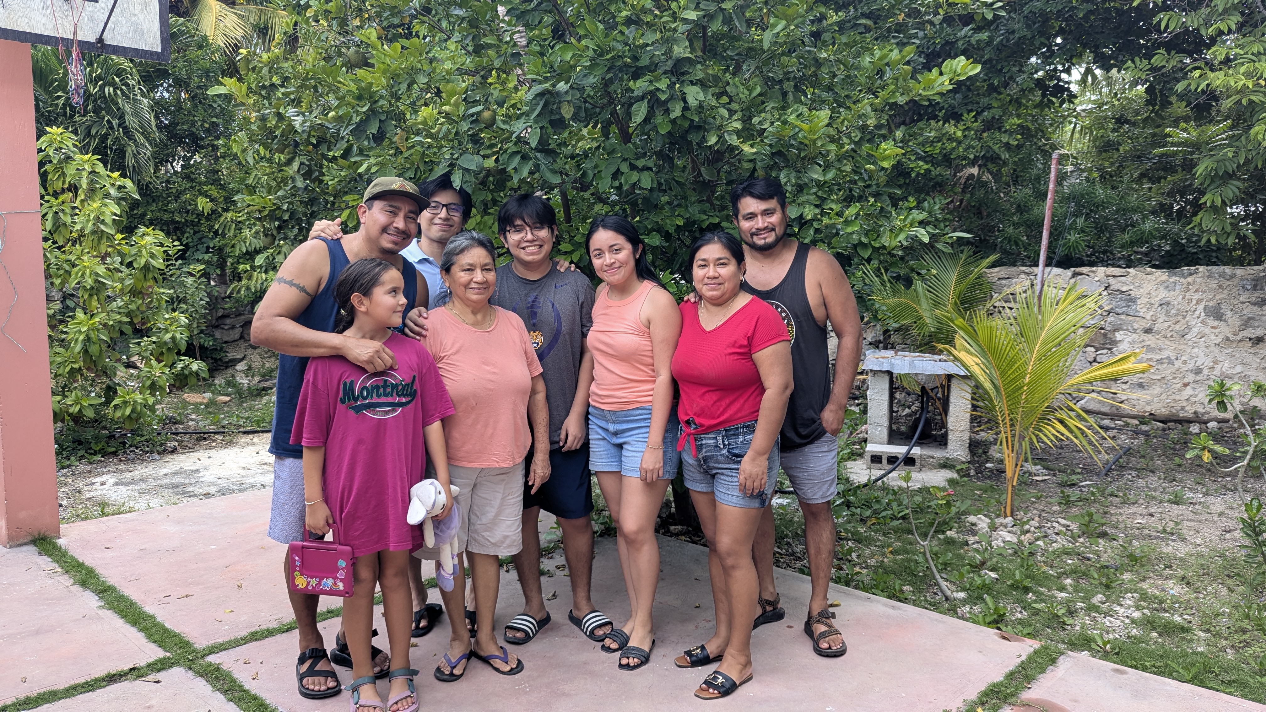 A group of eight people posing together outdoors, smiling in front of lush greenery. They are casually dressed and appear to be enjoying a gathering.